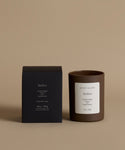 A candle in a dark brown glass jar with a white label next to its black packaging box, both featuring the brand name 'Jenii Kayne' and the scent 'Amber', set against a solid beige background.