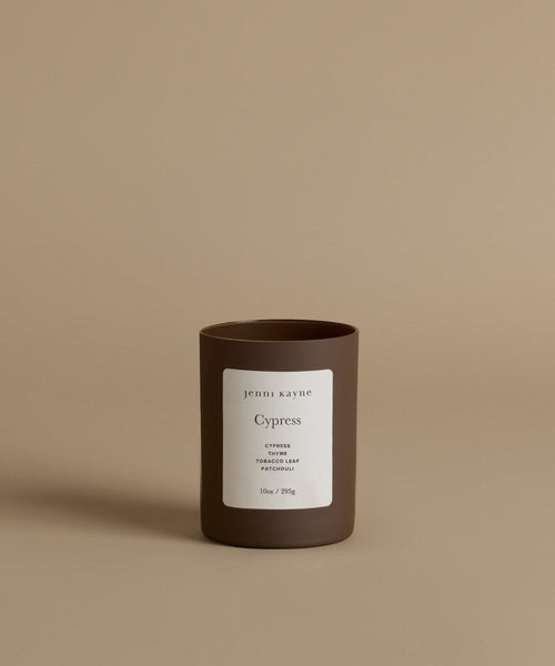 A glass candle in th scent Cypress in a brown vessel against a beige background.