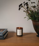 A glass candle on a wood table with flowers and books against a white wall.