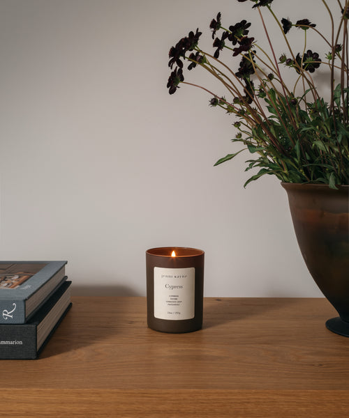 A glass candle on a wood table with flowers and books against a white wall.
