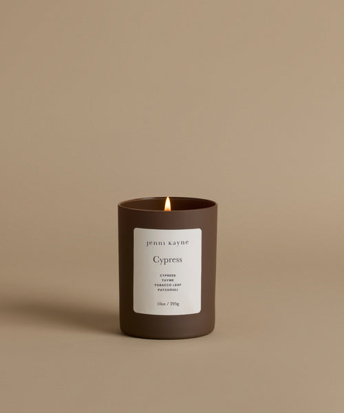 A lit candle in a matte dark brown glass jar with a white label reading 'jenni kayne' and 'Cypress', set against a solid warm beige background.