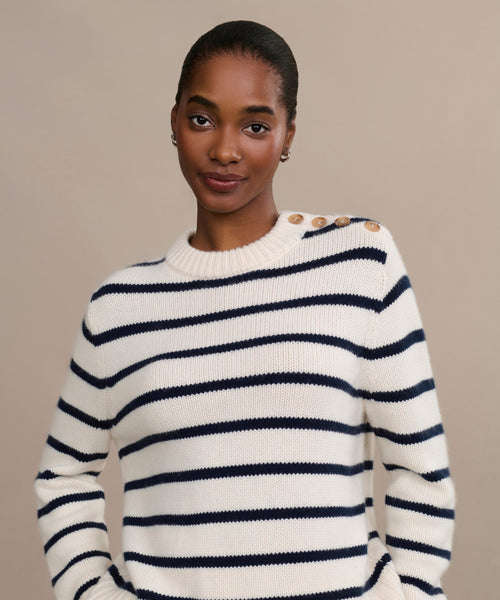 A woman smiles softly, hands in her pockets, wearing the Grace Crewneck—a luxury navy stripe knit sweater with button details on one shoulder—against a plain beige background.