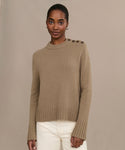 A woman models the Grace Crewneck, a luxurious knit tan sweater with shoulder buttons, paired with white pants against a simple beige background, gazing at the camera with a slight smile.