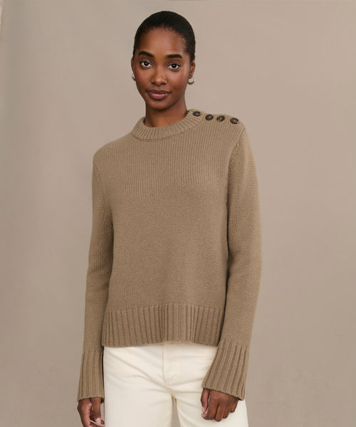 A woman models the Grace Crewneck, a luxurious knit tan sweater with shoulder buttons, paired with white pants against a simple beige background, gazing at the camera with a slight smile.