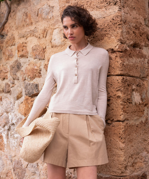 A woman with short curly hair stands against a stone wall, wearing the beige Grayson Pullover and tan shorts. She holds a woven straw hat in one hand and gazes slightly to the side with a neutral expression.