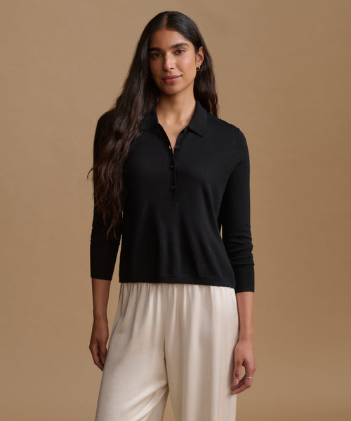 A woman modeling a black long-sleeve polo shirt with a partial button placket, paired with cream-colored pants, set against a solid warm beige studio background.