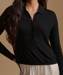 A woman wearing a black long-sleeve polo shirt with a buttoned collar, paired with light beige bottoms, set against a solid warm brown background.