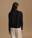 A woman viewed from the back wearing a black long-sleeve mock neck knit top and cream-colored trousers, set against a solid warm beige studio background.