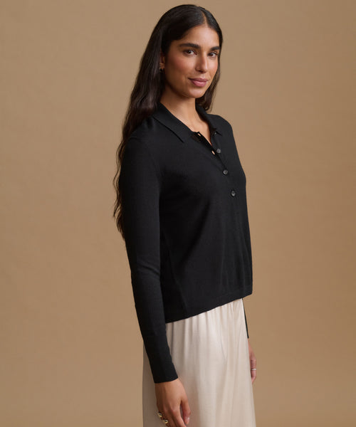 A woman modeling a black long-sleeve polo cardigan with a buttoned collar, paired with a cream-colored skirt, set against a solid warm beige studio background.