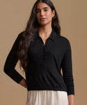 A woman modeling a black long-sleeve polo knit top with a buttoned placket, paired with a light cream skirt, against a solid warm beige studio background.