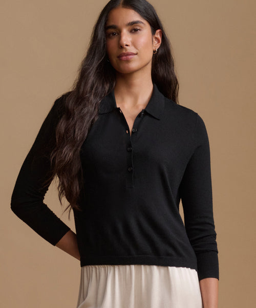 A woman modeling a black long-sleeve polo knit top with a buttoned placket, paired with a light cream skirt, against a solid warm beige studio background.