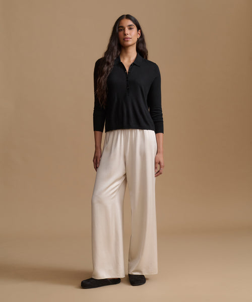 A woman stands against a warm beige studio background wearing a black long-sleeve polo-style top and wide-leg cream pants with black shoes.