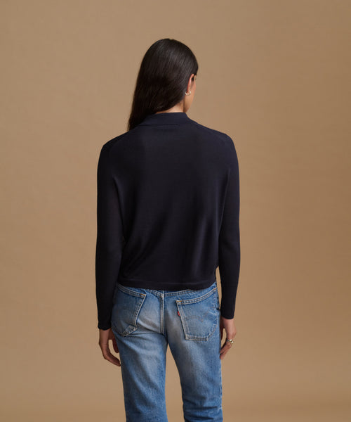 A woman viewed from the back wearing a dark navy long-sleeve knit polo-style top and light blue jeans, standing against a plain warm tan studio background.