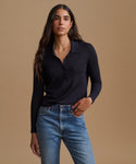 A woman modeling a dark navy long-sleeve polo shirt tucked into blue jeans, standing against a solid warm beige studio background.