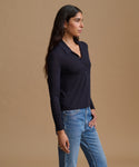 A woman wearing a dark navy long-sleeve polo shirt and light blue jeans, standing against a solid beige studio background.