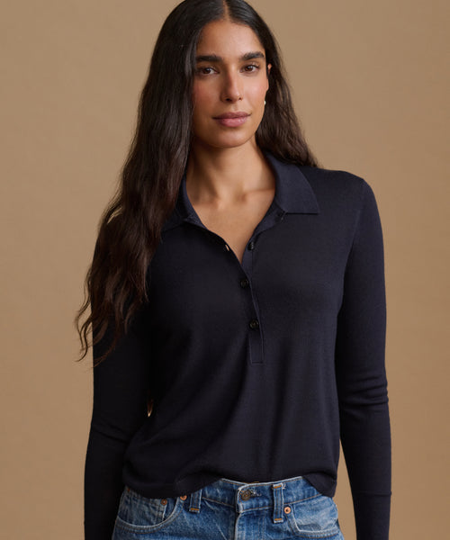 A woman modeling a dark navy long-sleeve polo shirt with a buttoned collar, paired with blue jeans, against a solid warm beige background.