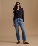 A woman modeling a dark navy long-sleeve polo shirt and blue denim jeans against a solid beige studio background.