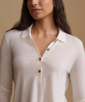 A close-up of a woman wearing a cream-colored knit polo shirt with brown buttons, set against a solid beige background.