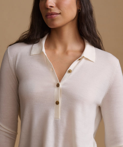 A close-up of a woman wearing a cream-colored knit polo shirt with brown buttons, set against a solid beige background.