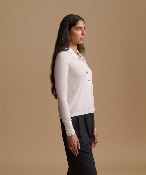 A woman in side profile wearing a cream long-sleeve button-front cardigan over a light beige collared shirt and black trousers, set against a solid warm beige studio background.