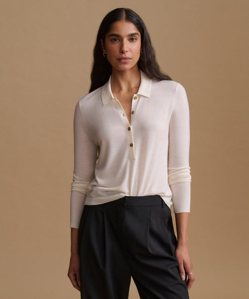 A woman modeling a cream-colored long-sleeve knit polo shirt with a buttoned collar, paired with black trousers, against a solid beige studio background.