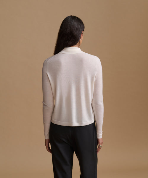 A woman viewed from the back wearing a light cream mock-neck knit sweater and black trousers, standing against a solid tan studio background.