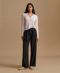 A woman stands against a warm beige studio background wearing a light cream long-sleeve button-front top, black wide-leg pants, and tan slip-on shoes.