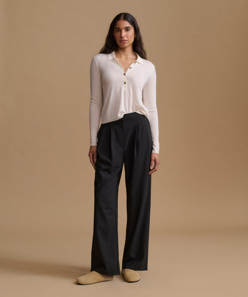 A woman stands against a warm beige studio background wearing a light cream long-sleeve button-front top, black wide-leg pants, and tan slip-on shoes.