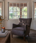 A gray upholstered wingback armchair with button tufting on the back, placed in a bright room with large windows, wood flooring, a wooden table with books and a candle, and a woven rug underneath.