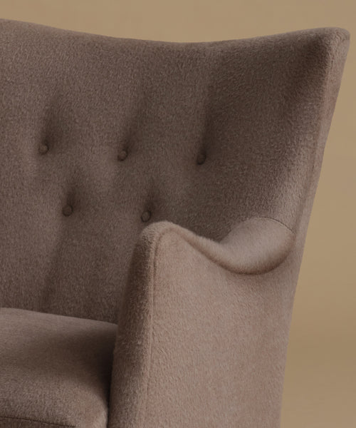 Close-up of a tufted armchair upholstered in a textured taupe fabric, showing the button-tufted backrest and curved arm against a warm beige background.