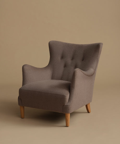 A single upholstered wingback armchair with a high tufted back, curved arms, and tapered wooden legs, shown on a plain warm beige studio background.