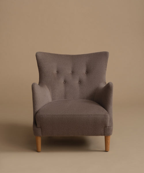 A single upholstered armchair with a high tufted back, curved arms, and light wood legs, shown against a plain warm beige studio background.