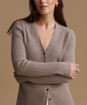 A woman wearing a ribbed knit cardigan sweater in taupe brown, shown from the chest up against a solid warm beige background.