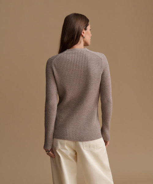 A woman shown from the back wearing a taupe ribbed knit sweater and cream wide-leg trousers, set against a solid warm beige studio background.