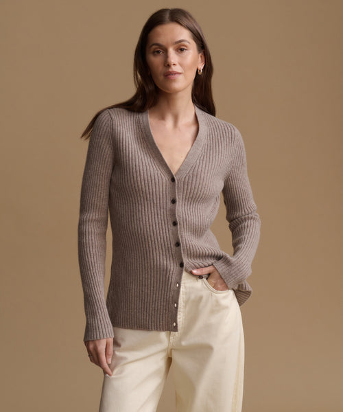 A woman modeling a taupe ribbed knit cardigan sweater with dark buttons, paired with cream pants, against a solid warm beige studio background.