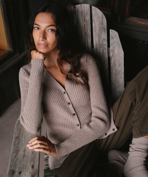 A woman wearing a taupe ribbed button-front cardigan sweater sits in a rustic wooden Adirondack chair in a dimly lit interior setting.