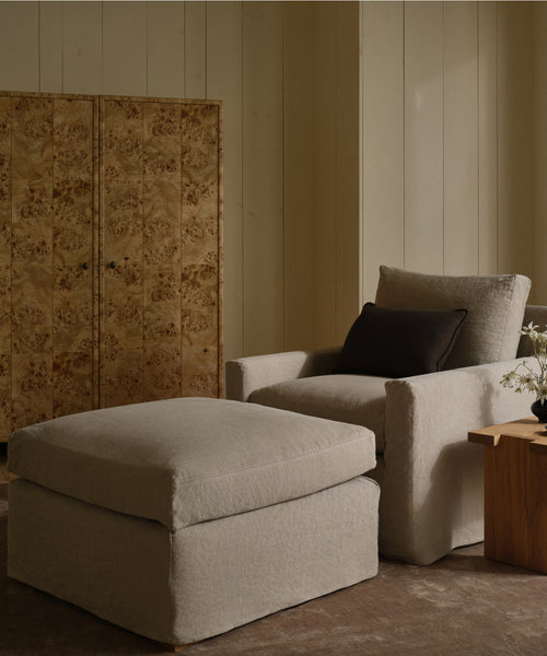 A cozy living space with a light beige armchair, Harbor Ottoman by Jenni Kayne Furniture, dark accent pillow, wooden side table with a vase of flowers, and a decorative wood-paneled screen against cream-colored walls.