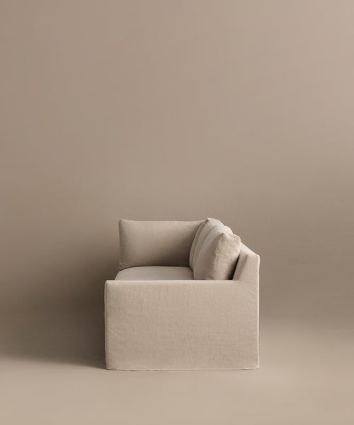 The Harbor Sectional, a minimalist Belgian flax linen sofa with clean lines and plush cushions, is set against a matching beige background and photographed from the side.