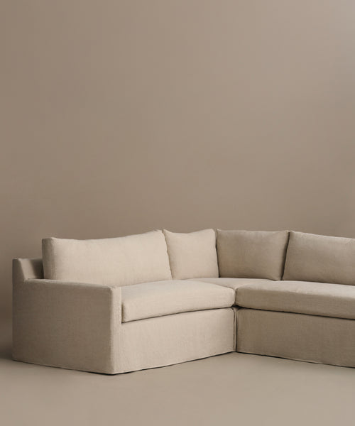 The Harbor Sectional in beige Belgian flax linen features clean lines and plush cushions, positioned against a light brown wall and floor—a cozy piece inspired by Jenni Kayne Furniture.