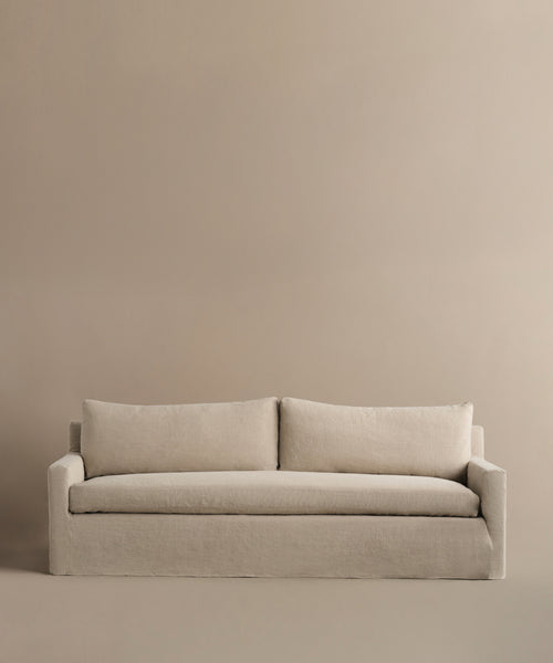 The Harbor Sofa features a modern minimalist design with clean lines, two large back cushions, and linen upholstery in beige, perfectly complemented by a matching beige background.