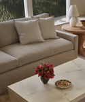 A modern living room with a linen-upholstered beige Harbor Sofa and two matching cushions, a round wooden side table with a white lamp, and a marble coffee table with red flowers. Sunlight pours in through large windows.