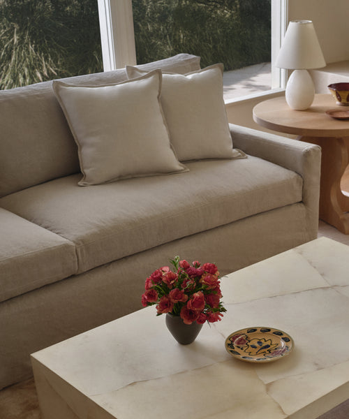 A modern living room with a linen-upholstered beige Harbor Sofa and two matching cushions, a round wooden side table with a white lamp, and a marble coffee table with red flowers. Sunlight pours in through large windows.
