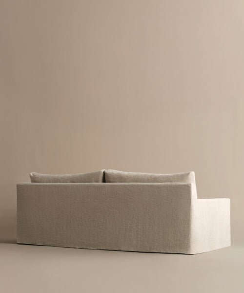 The Harbor Sofa features a modern, minimalist design with linen upholstery and two back cushions, set against a light beige background. Its clean lines and neutral color offer a calm, contemporary vibe.