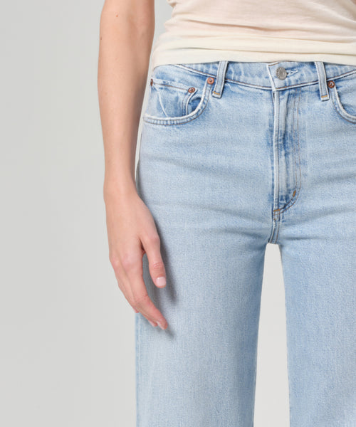 A person wears the Harper Jean in light blue organic cotton with a mid-rise, wide-leg fit and a beige top, shown waist to mid-thigh, left arm relaxed by their side against a plain light background.