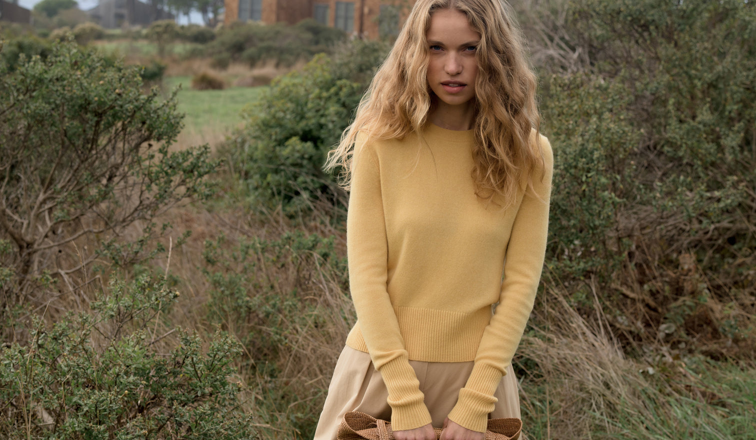 Woman in a yellow sweater standing in a natural setting with greenery.