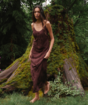 A woman stands barefoot outdoors in a mossy forest setting, wearing a dark brown satin slip dress with a subtle tonal floral pattern.