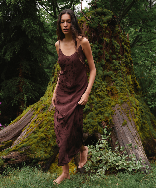 A woman stands barefoot outdoors in a mossy forest setting, wearing a dark brown satin slip dress with a subtle tonal floral pattern.