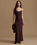 A woman stands against a warm beige studio background wearing a long, dark plum-brown satin slip dress with thin straps and holding a matching clutch bag.