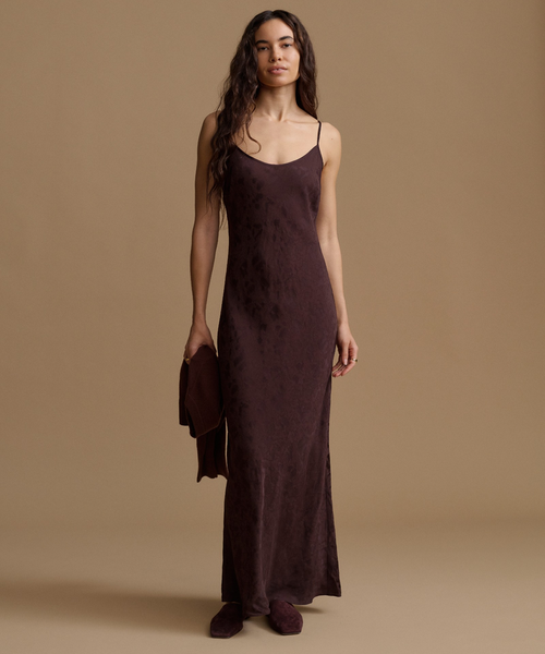 A woman stands against a warm beige studio background wearing a long, dark plum-brown satin slip dress with thin straps and holding a matching clutch bag.