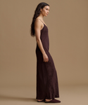 A woman in profile wearing a long, dark brown satin slip dress and matching flat shoes, standing against a solid beige studio background.